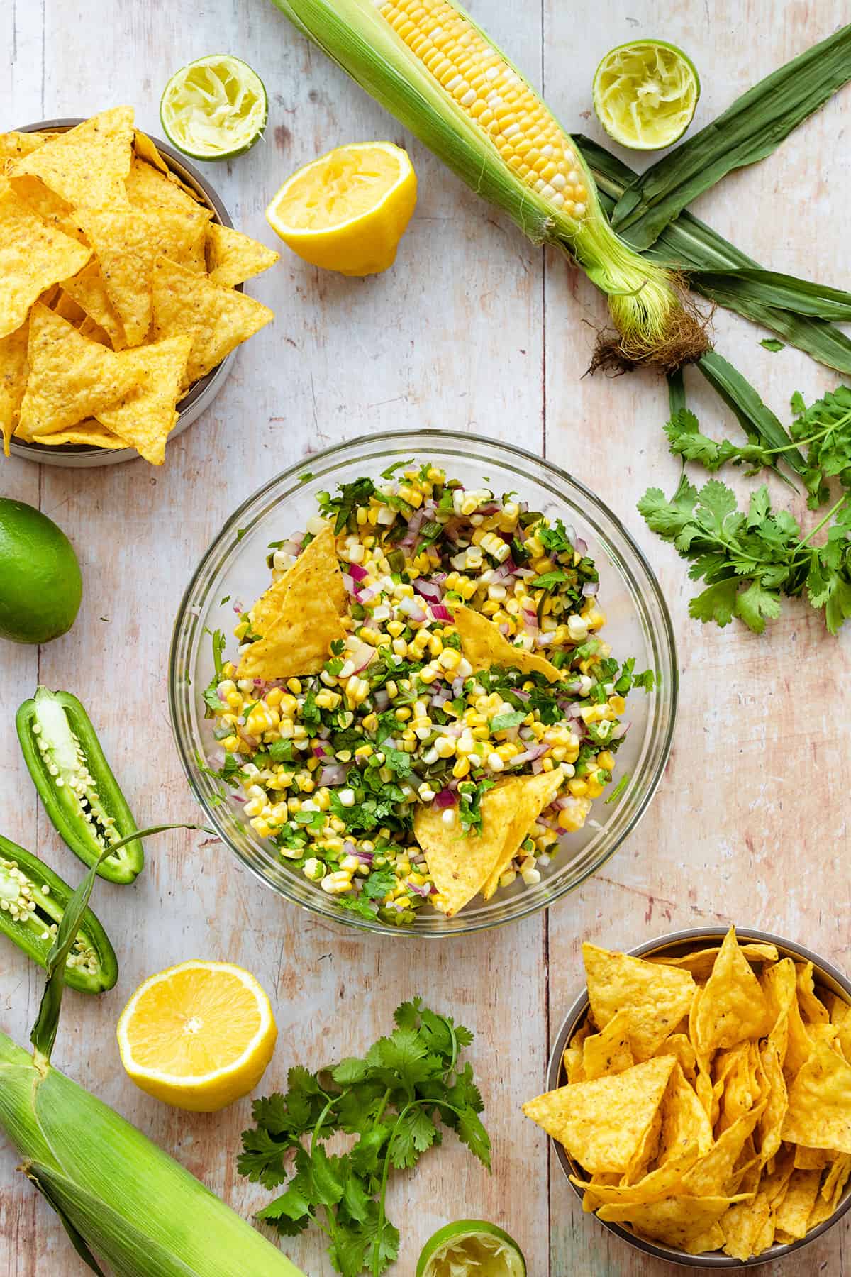 Corn salsa in a glass bowl with tortilla chips dipped in. There are more tortilla chips, fresh cilantro, corn, and jalapenos around. All on a light wooden background.