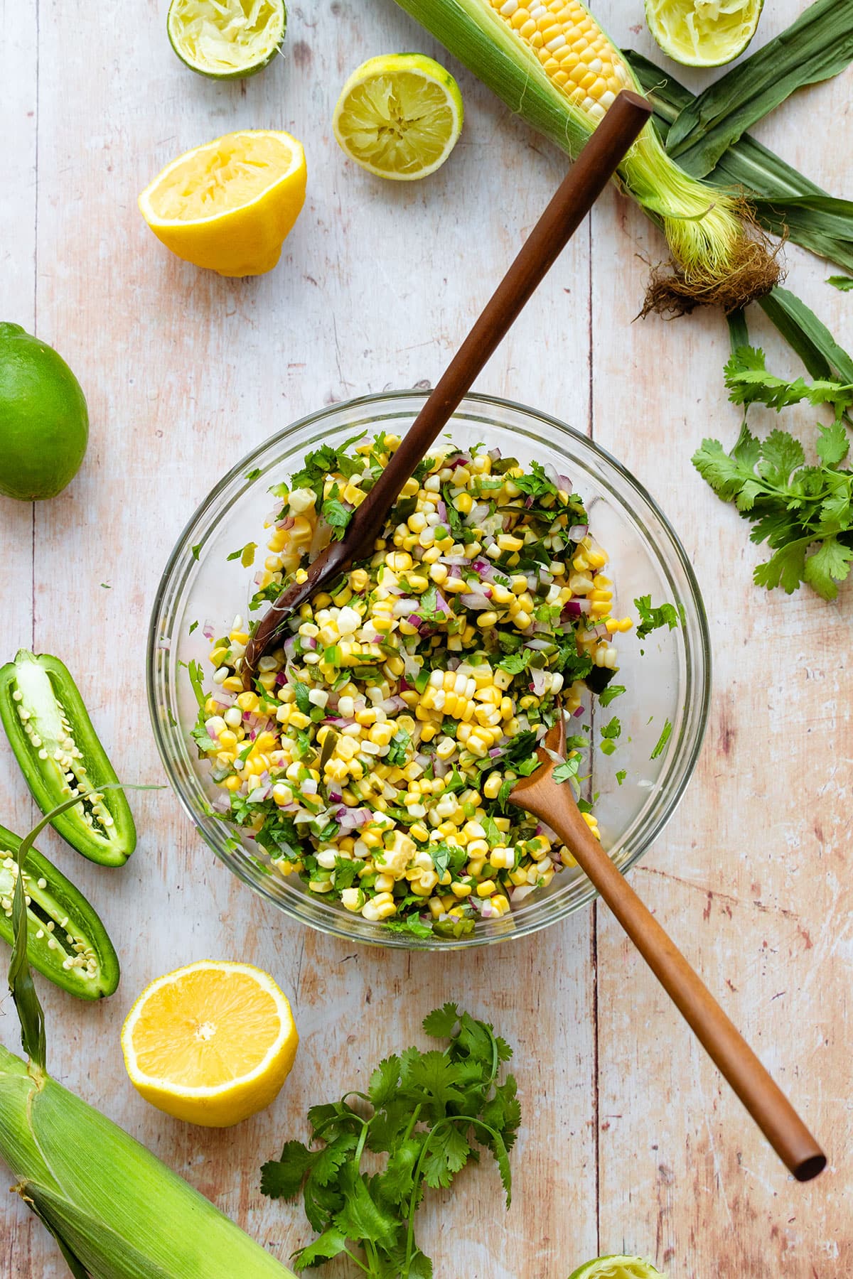 Corn salsa in a glass bowl with two wooden mixing spoons in it. There are more tortilla chips, fresh cilantro, corn, and jalapenos around. All on a light wooden background.