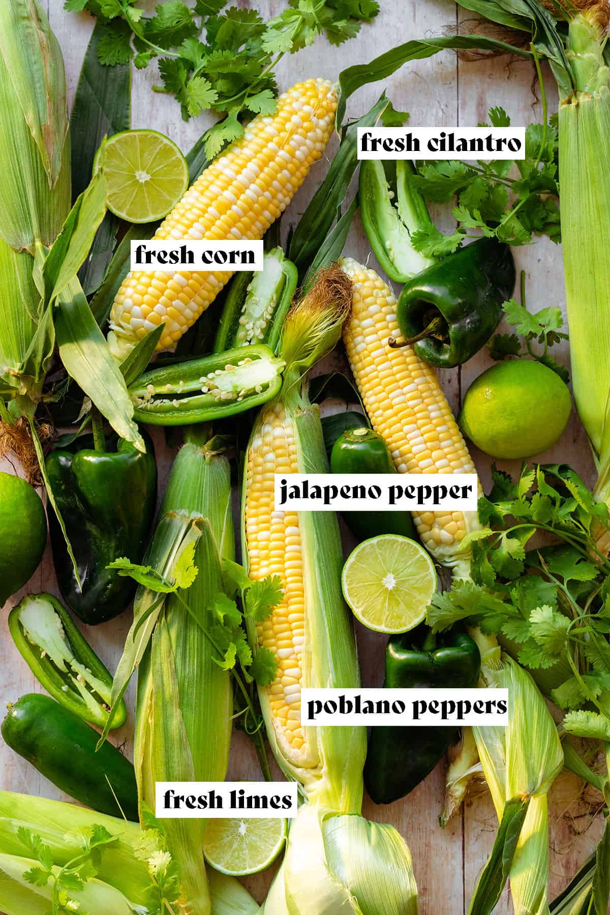 Ingredients for Roasted Chili Corn Salsa all laid out on a light wooden background. There's corn, cilantro, jalapeno pepers, poblano peppers, and fresh lime.