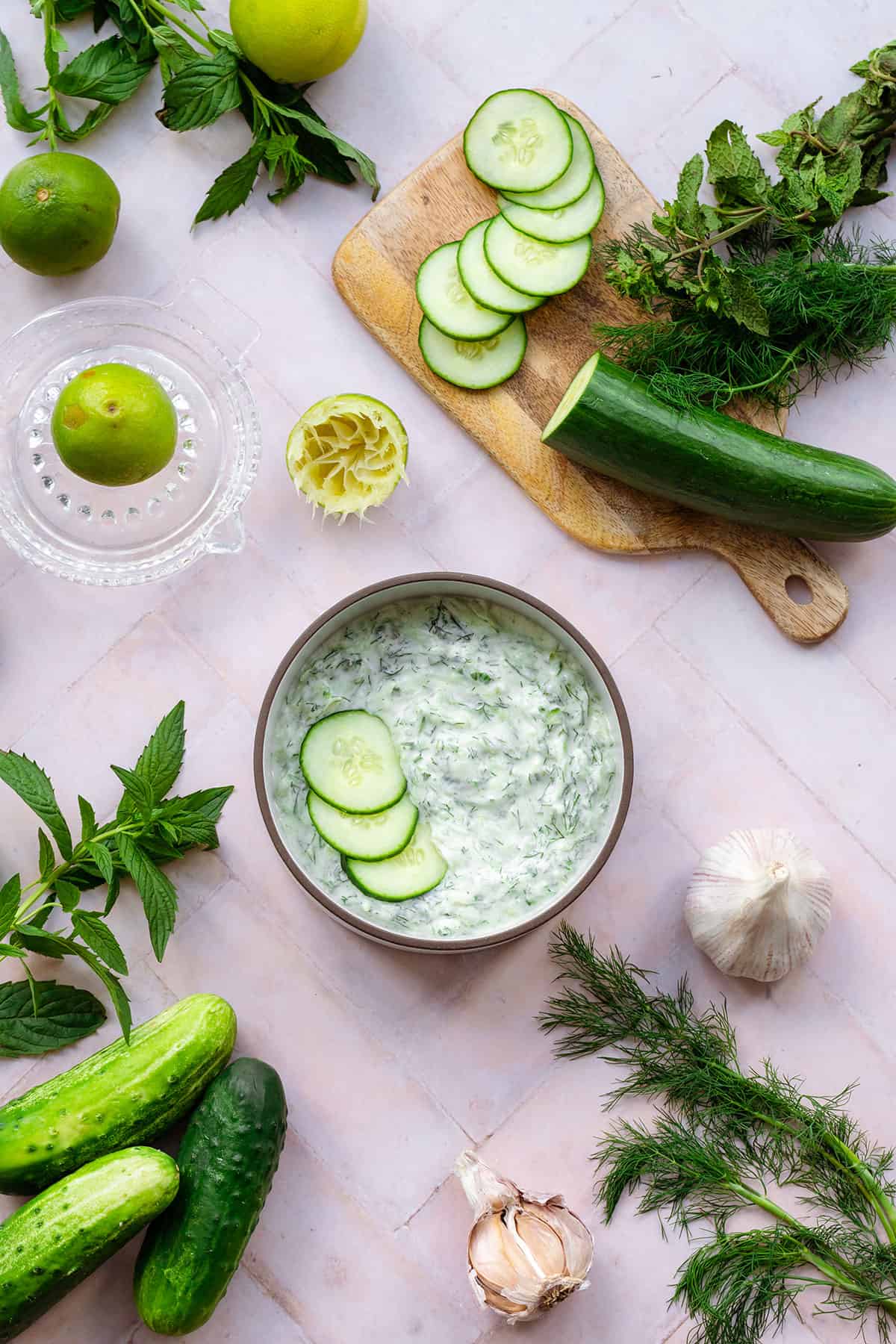 Tzatziki Sauce in a white bowl with a brown rim garnished with three cucumber slices. On a pink tile background with cucumber, limes, garlic, mint, and dill all around.