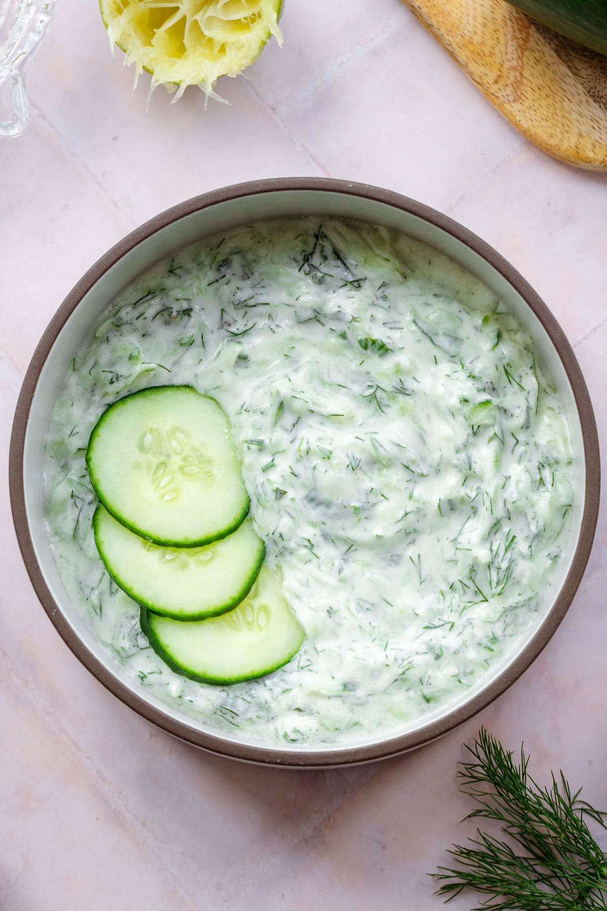 Tzatziki Sauce in a white bowl with a brown rim garnished with three cucumber slices. On a pink tile background with cucumber, a lime, and dill around.