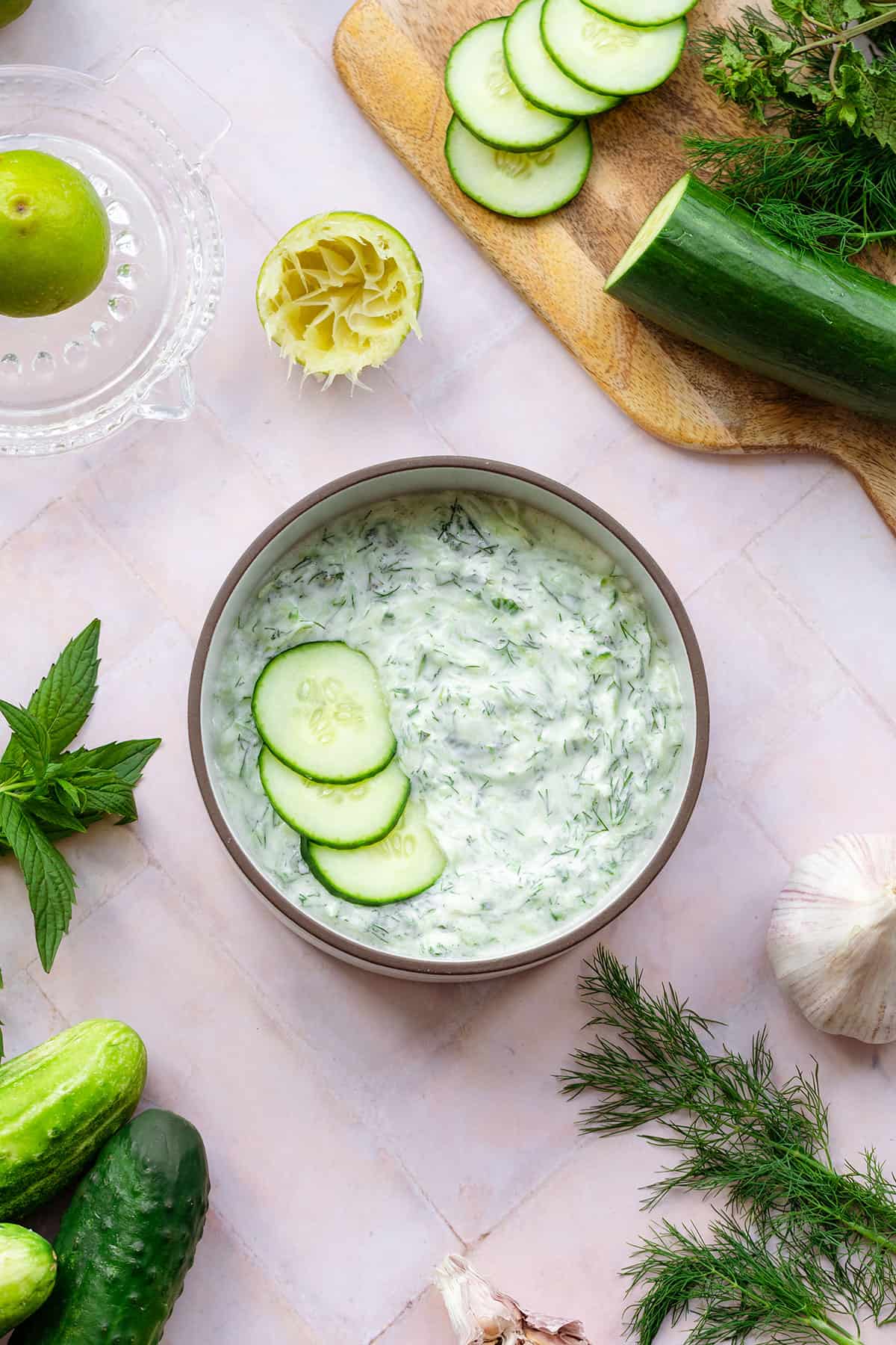 Tzatziki Sauce in a white bowl with a brown rim garnished with three cucumber slices. On a pink tile background with cucumber, limes, garlic, mint, and dill all around.