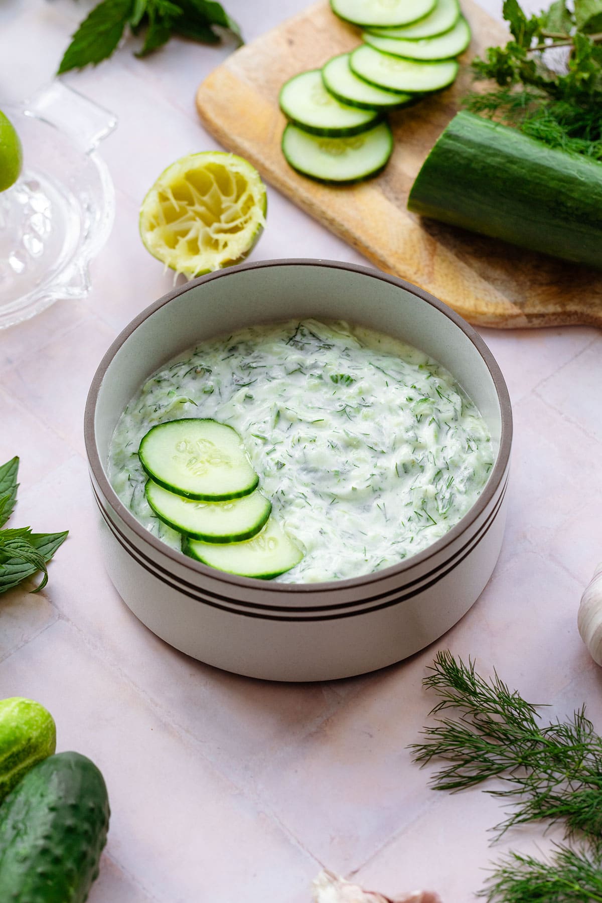 Tzatziki Sauce in a white bowl with a brown rim garnished with three cucumber slices. On a pink tile background with cucumber, limes, garlic, mint, and dill all around.