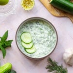 Tzatziki Sauce in a white bowl with a brown rim garnished with three cucumber slices. On a pink tile background with cucumber, limes, garlic, mint, and dill all around.