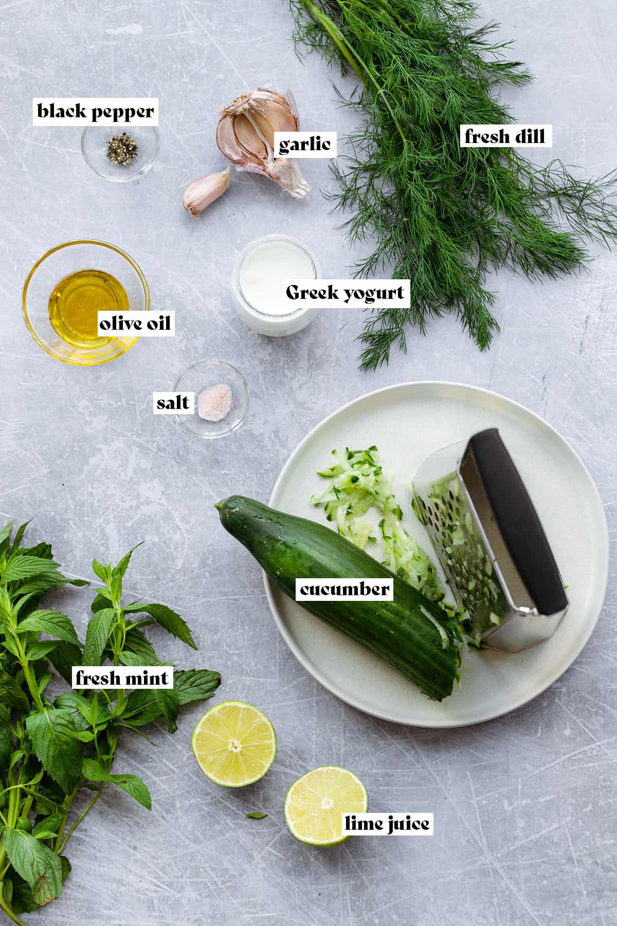 A photo of all the tzatziki ingredients laid out on a metal background. Fresh dill, mint, cucumber, limes, olive oil, greek yogurt, garlic, black pepper, and salt.