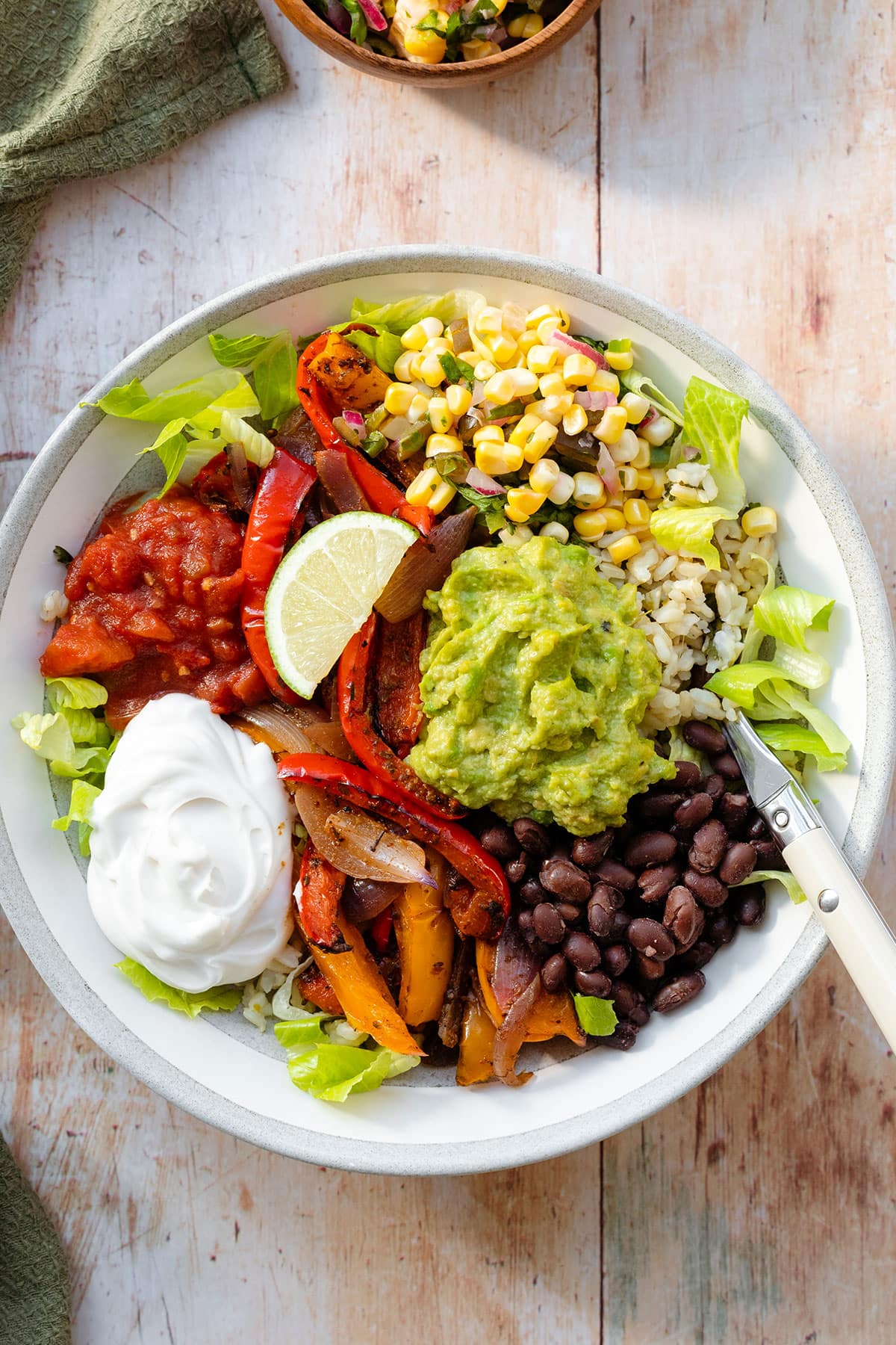 A photo of veggie burrito bowl in a grey bowl with a white rim on a light wooden background with a green kitchen towel on the left. In the bowl there is brown rice, corn salsa, tomato salsa, sour cream, guacamole, black beans, and roasted peppers.