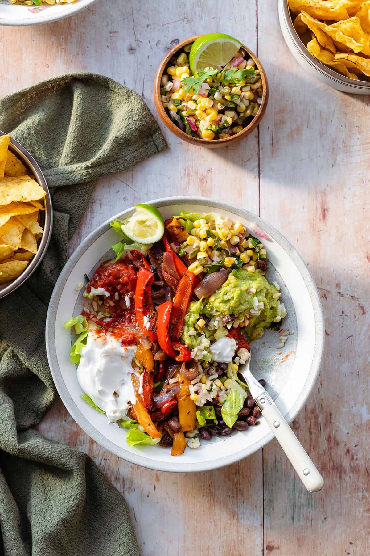 A photo of veggie burrito bowls in a grey bowls with a white rim on a light wooden background with a green kitchen towel on the left and a small bowl with corn salsa above the bowl in the middle. In the bowl there is brown rice, corn salsa, tomato salsa, sour cream, guacamole, black beans, and roasted peppers.