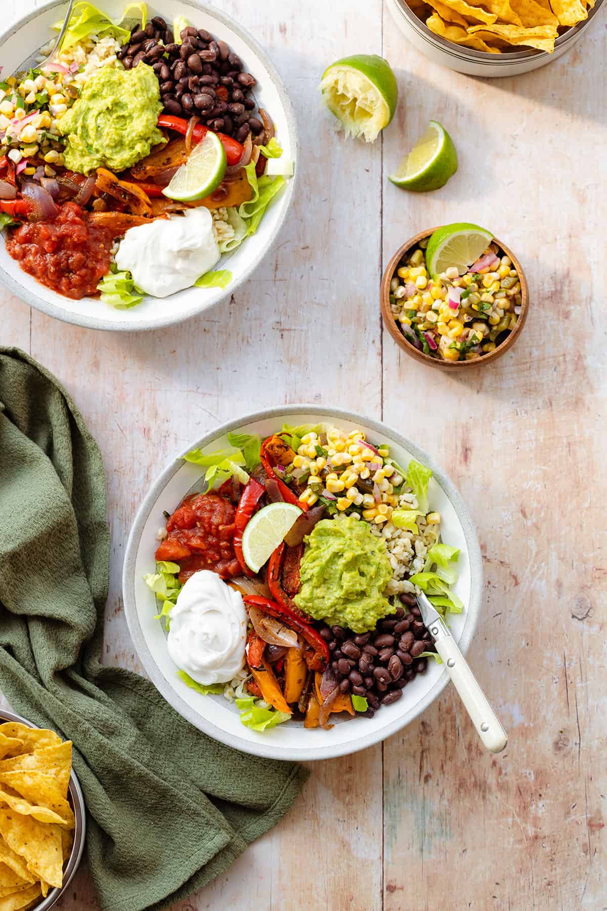 A photo of veggie burrito bowl in a grey bowl with a white rim on a light wooden background with a green kitchen towel on the left. In the bowl there is brown rice, corn salsa, tomato salsa, sour cream, guacamole, black beans, and roasted peppers.