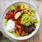 A photo of veggie burrito bowl in a grey bowl with a white rim on a light wooden background. In the bowl there is brown rice, corn salsa, tomato salsa, sour cream, guacamole, black beans, and roasted peppers.