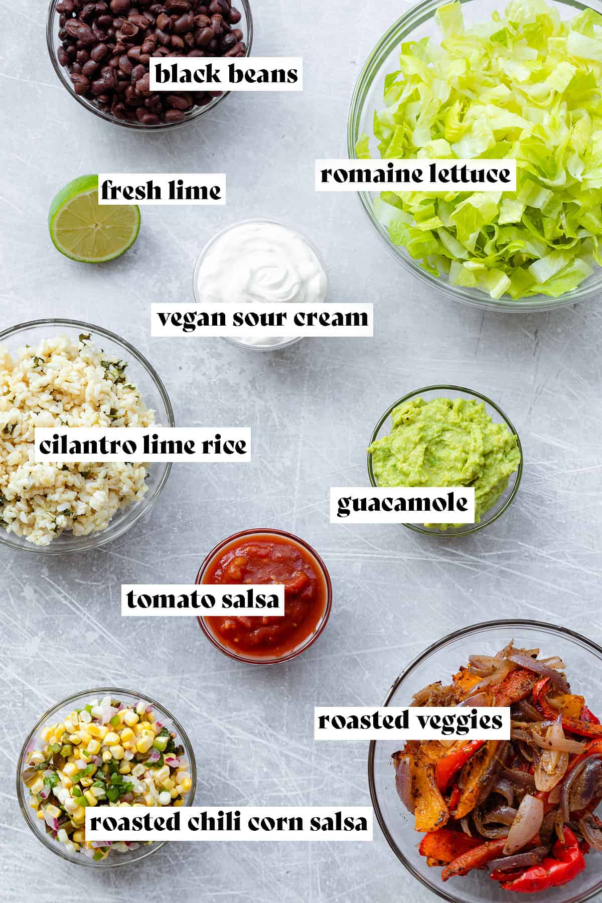 Ingredients in glass bowls on a steel background. Romaine lettuce, black beans, vegan sour cream, guacamole, cilantro lime rice, tomato salsa, roasted chili corn salsa, and roasted veggies.