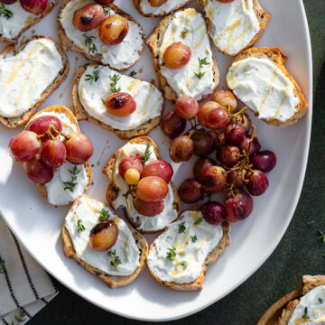 Crostini with goat cheese and roasted grapes on a white plate on a dark green background.