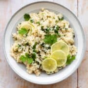 A photo of brown rice with cilantro and lime, decorated with lime slices. In a grey bowl with a white rim on a light wooden background. Juiced lime and fresh cilantro in the left top corner.