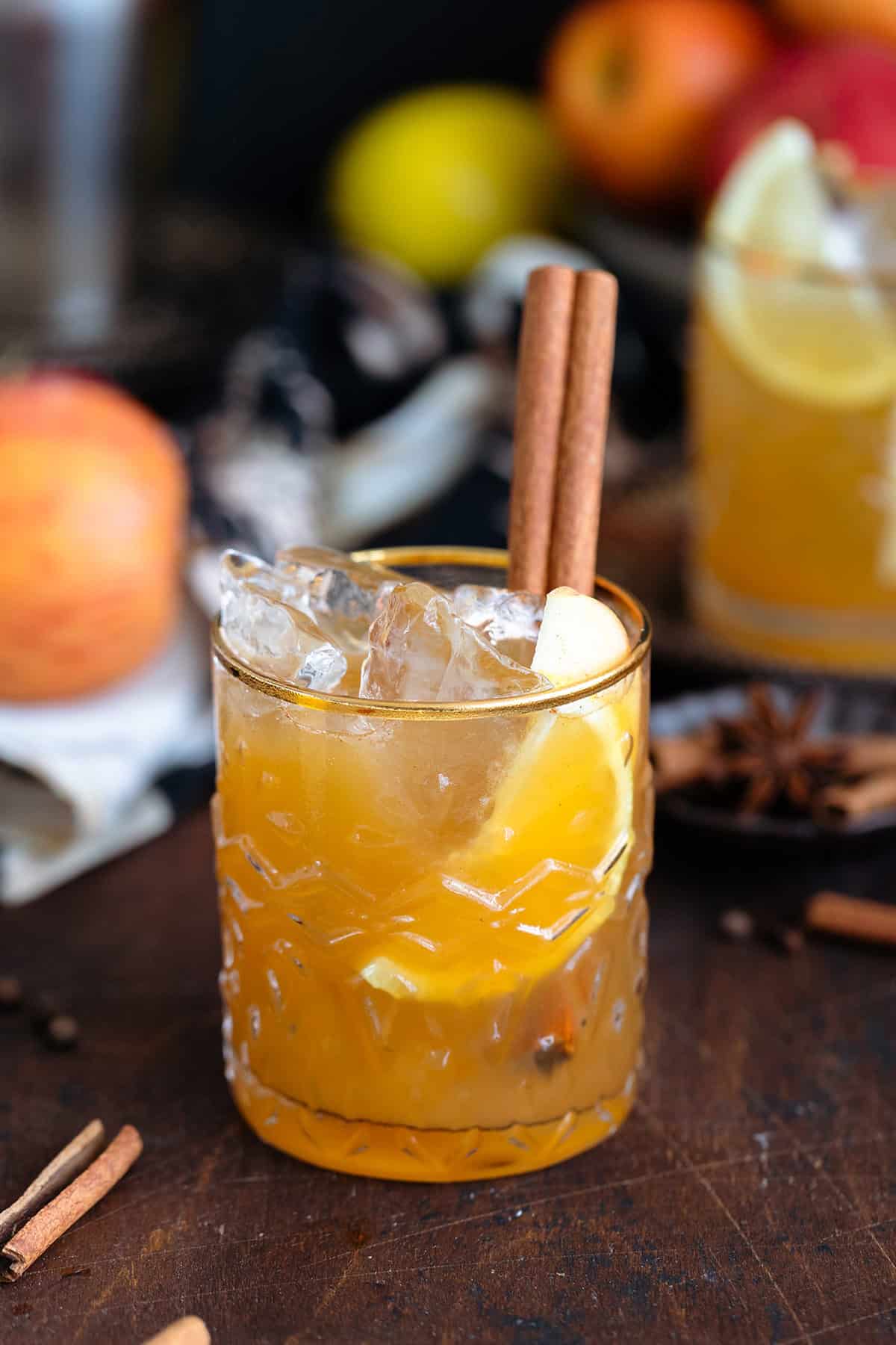 Bourbon cocktail in a glass with a gold rim and garnished with a cinnamon stick on a dark wooden background.