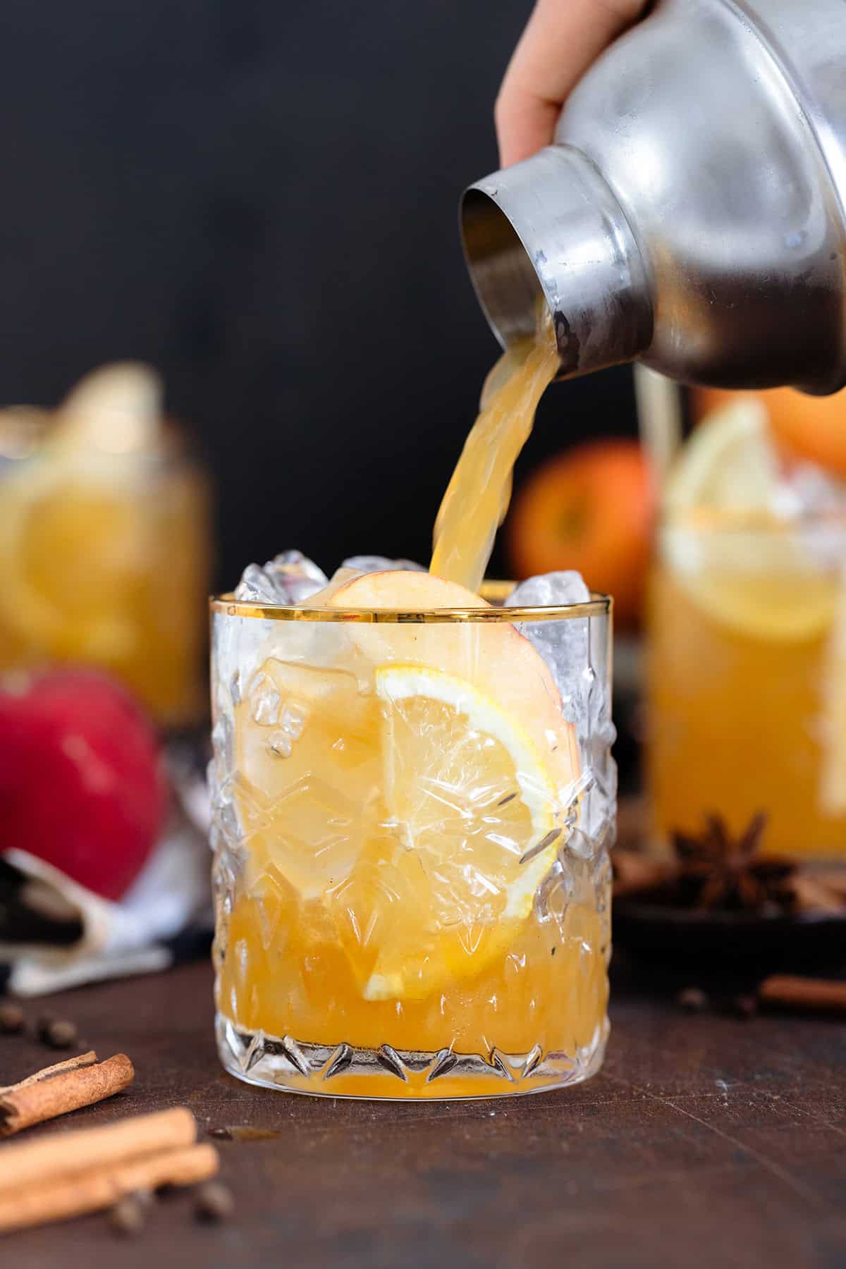 Bourbon cocktail being poured over ice into a glass with a gold rim on a dark wooden background.