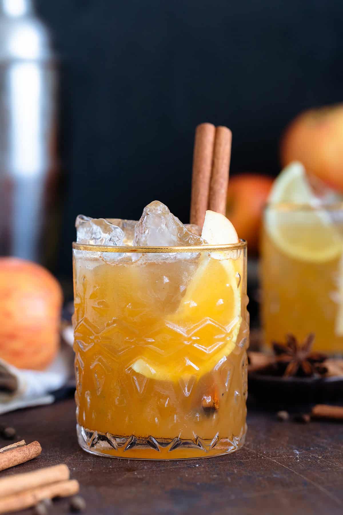 Bourbon cocktail in a glass with a gold rim and garnished with a cinnamon stick on a dark wooden background.