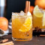 Bourbon cocktail in a glass with a gold rim and garnished with a cinnamon stick on a dark wooden background.