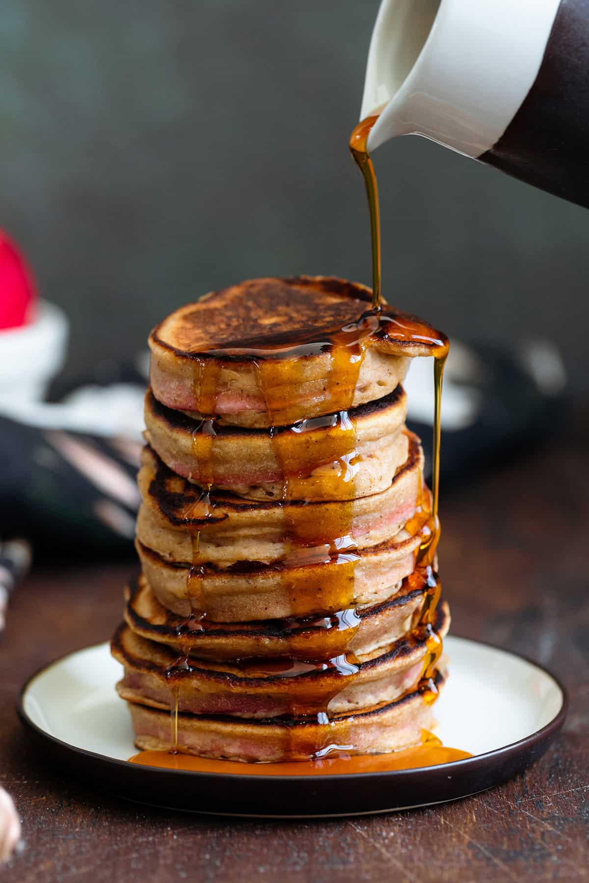 Maple syrup being poured over a stack of pancakes on a small plate on a dark wooden background.