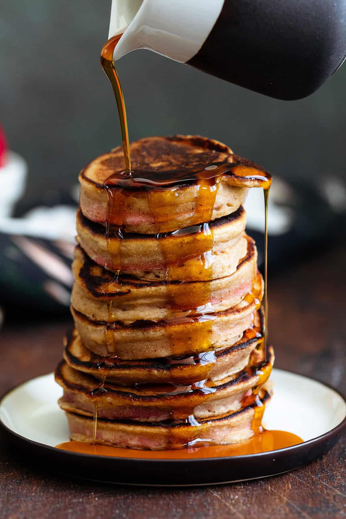 Maple syrup being poured over a stack of pancakes on a small plate on a dark wooden background.