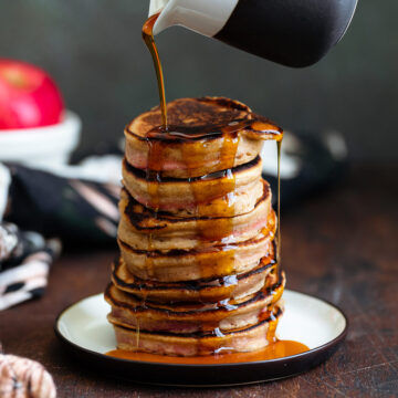 Maple syrup being poured over a stack of pancakes on a small plate on a dark wooden background.