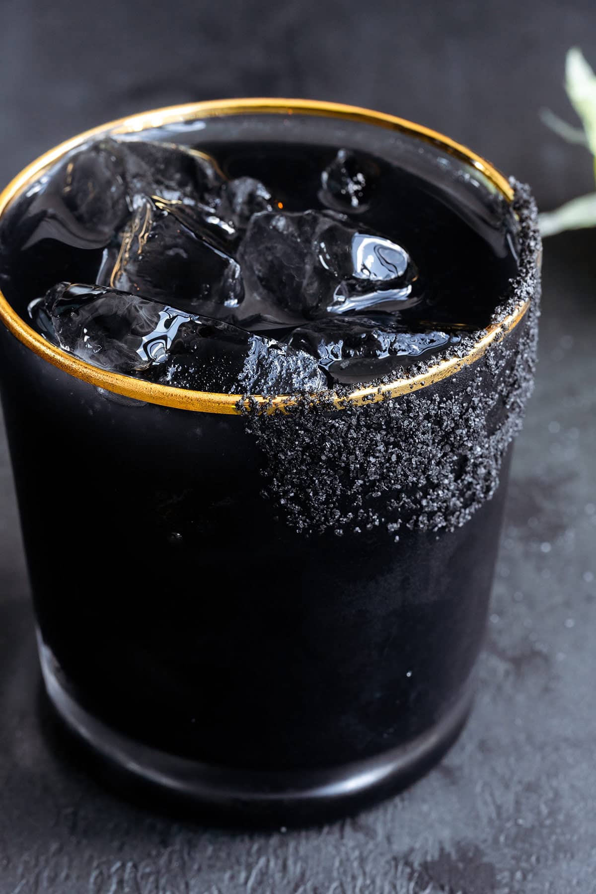 Black margarita with black salt rim on a black background with limes in the background.