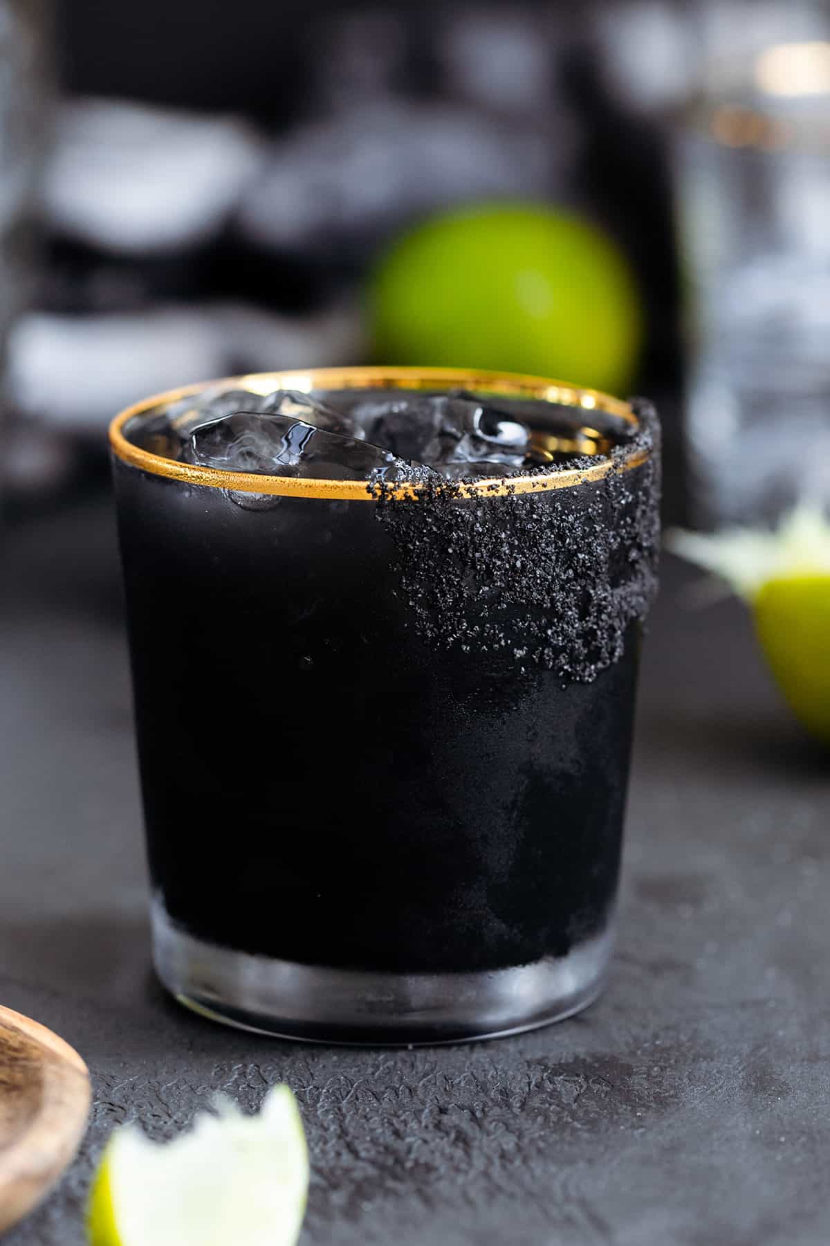 Black margarita with black salt rim on a black background with limes in the background.