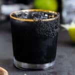 Black margarita with black salt rim on a black background with limes in the background.