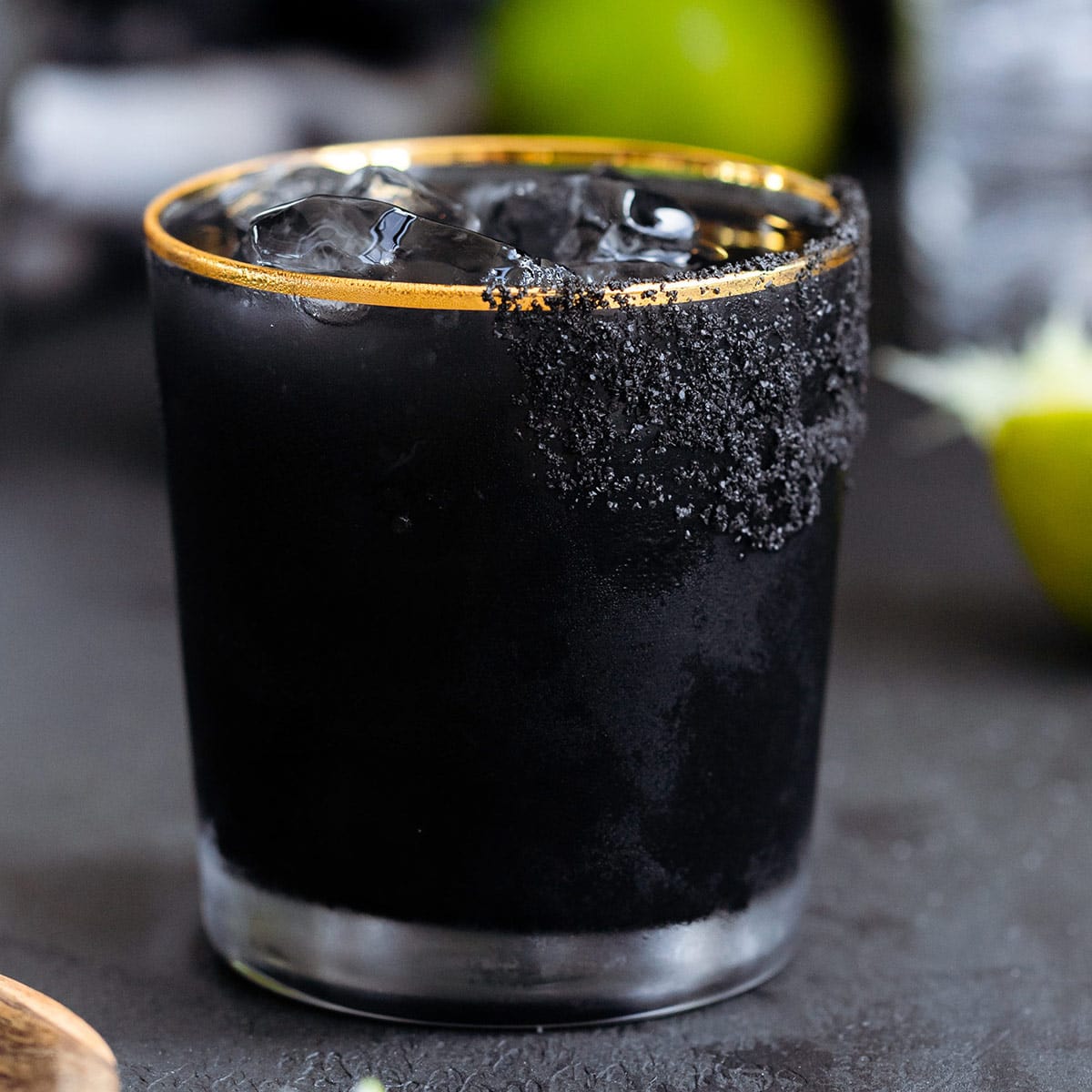 Black margarita with black salt rim on a black background with limes in the background.