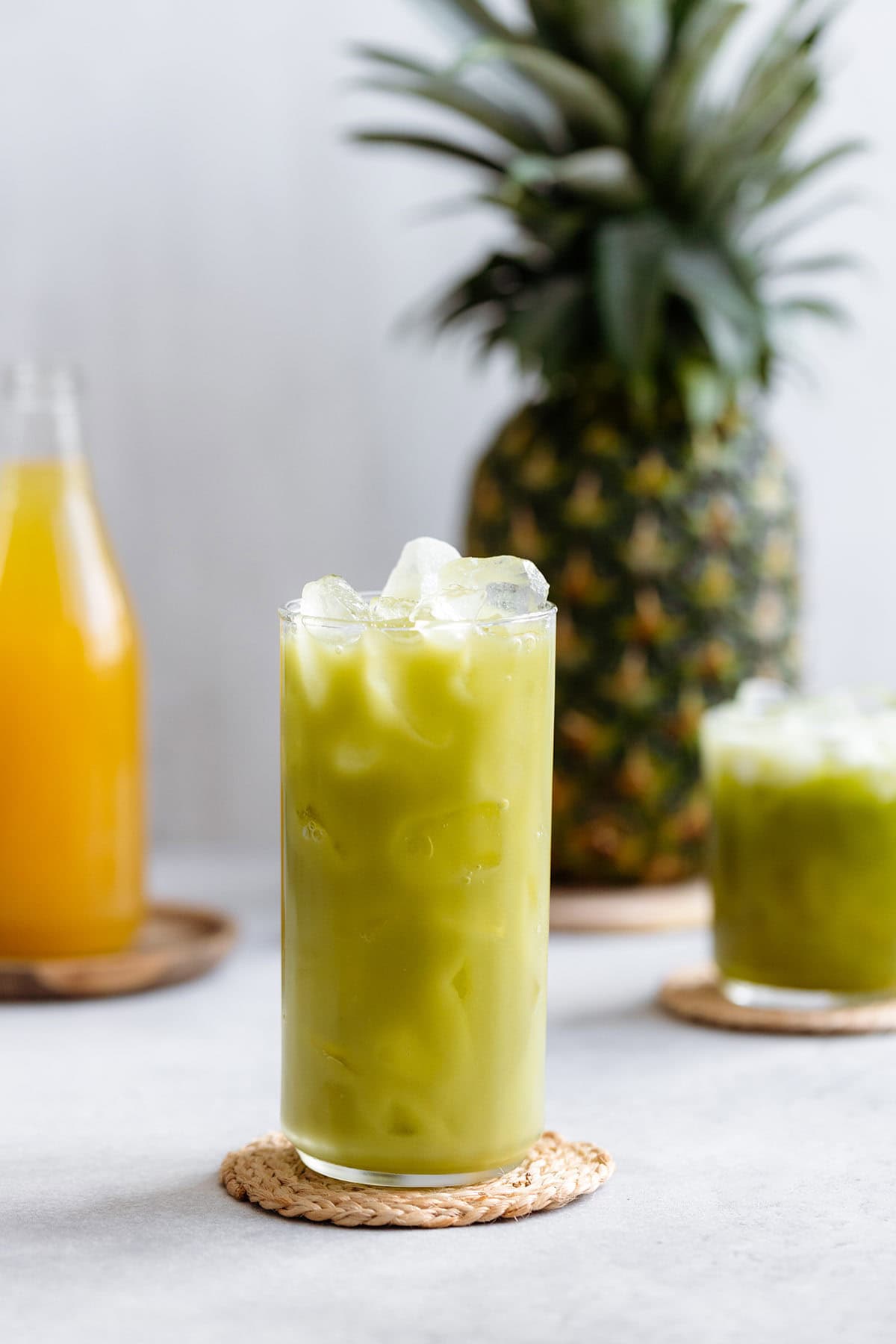 Pineapple drink in a tall glass served over ice on a white background with pineapple in the background.