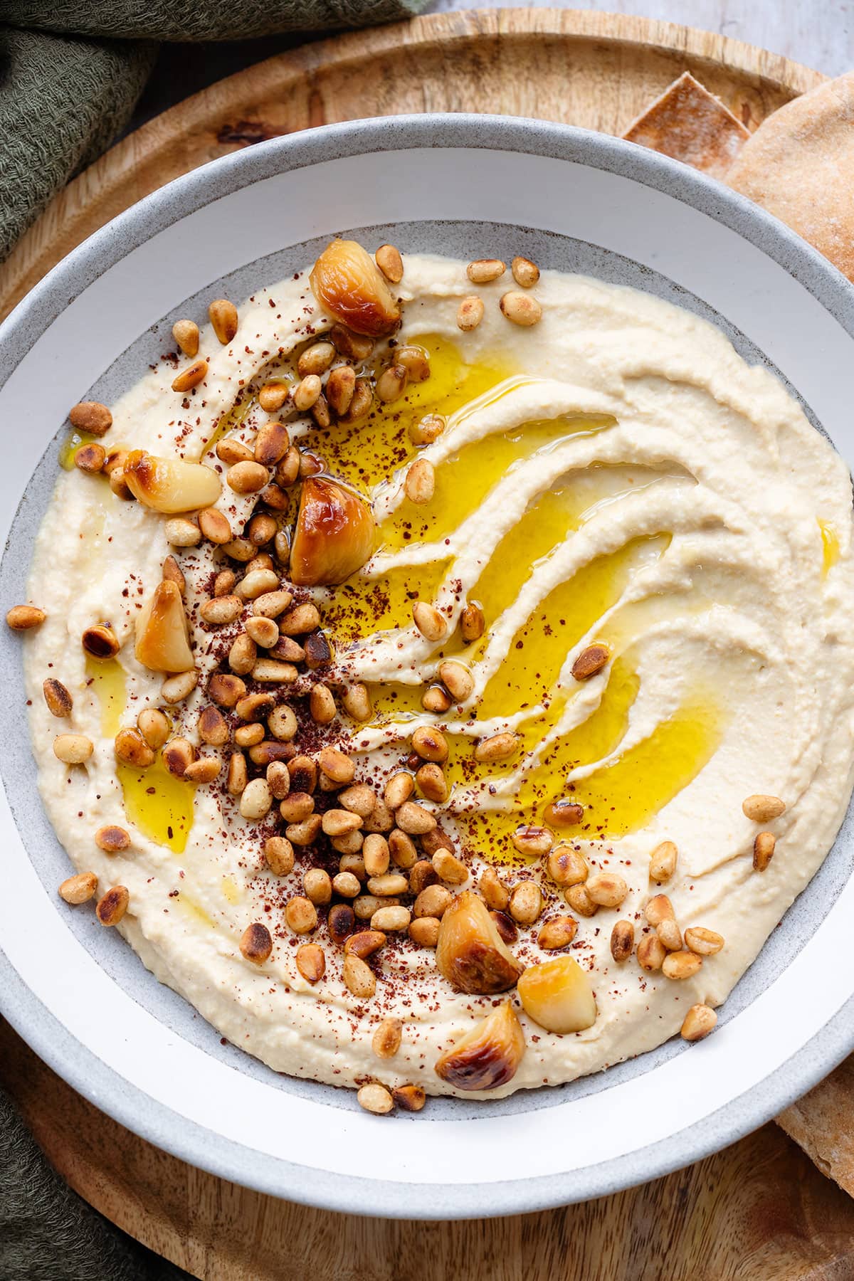 A close up of hummus in a shallow grey bowl topped with pine nuts, roasted garlic, and olive oil.