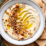 Hummus in a shallow grey bowl topped with pine nuts, roasted garlic, and olive oil.