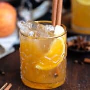 Bourbon cocktail in a glass with a gold rim and garnished with a cinnamon stick on a dark wooden background.