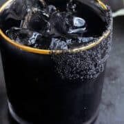 Black margarita with black salt rim on a black background with limes in the background.