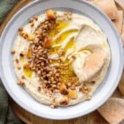 Hummus in a shallow grey bowl topped with pine nuts, roasted garlic, and olive oil with a pita dipped in.