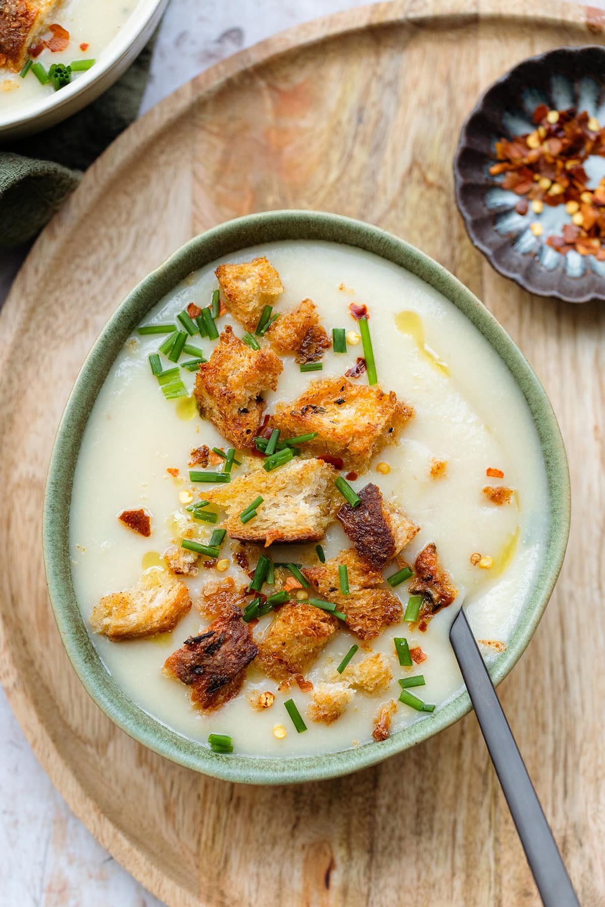 Cauliflower soup topped with croutons, fresh chives, and chili flakes in a green bowl on a wooden decorative plate.
