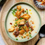 Cauliflower soup topped with croutons and fresh herbs in a green bowl on a wooden decorative plate.