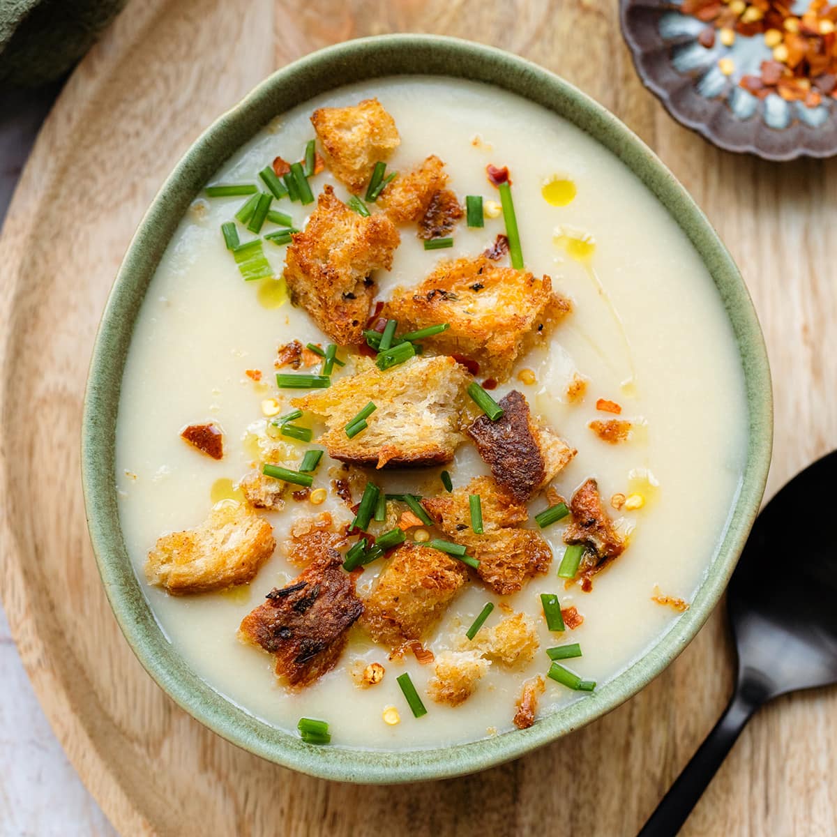 Cauliflower soup topped with croutons and fresh herbs in a green bowl on a wooden decorative plate.