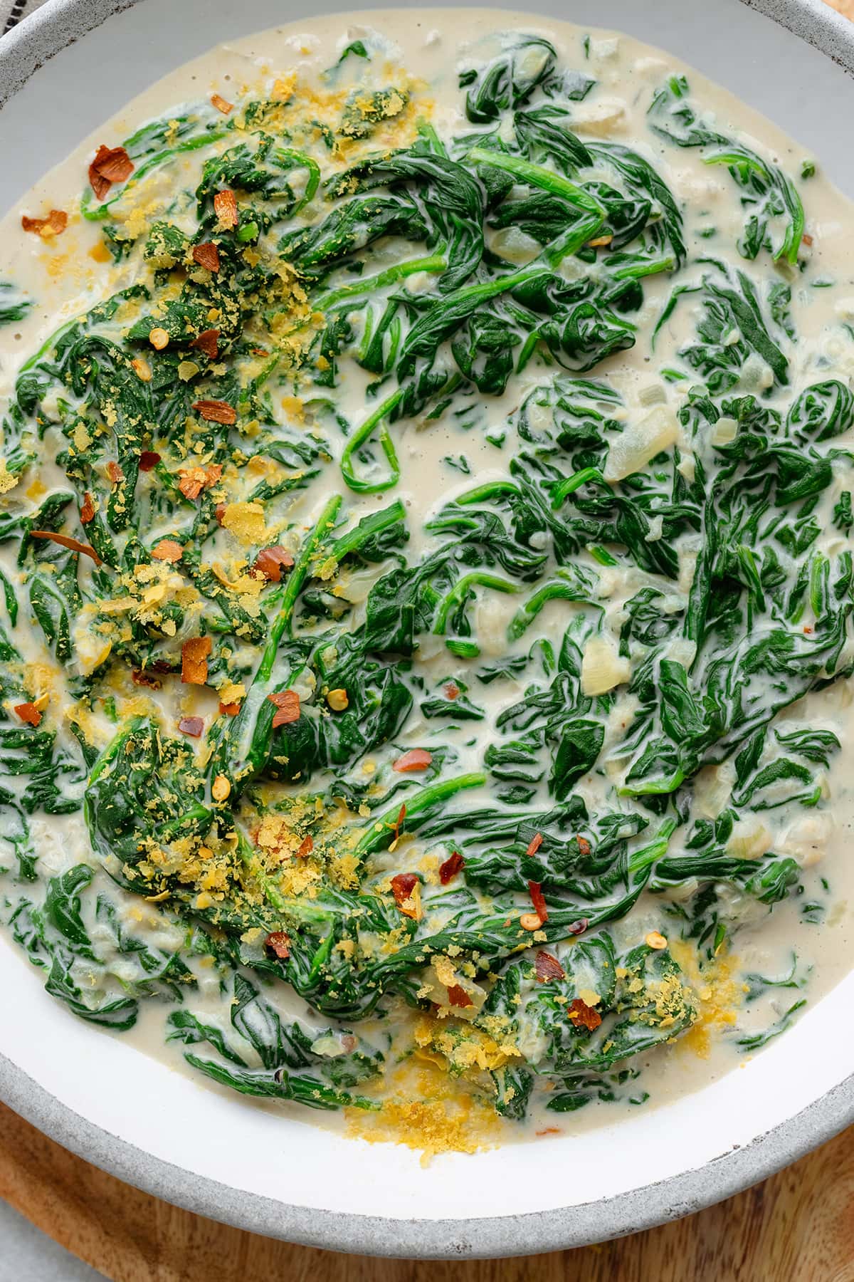 A close up of Creamed spinach on a grey ceramic bowl sprinkled with nutritional yeast and chili flakes.