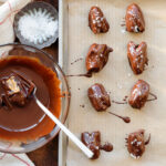 Dates covered in dark chocolate and sprinkled with flaky sea salt on a baking sheet.