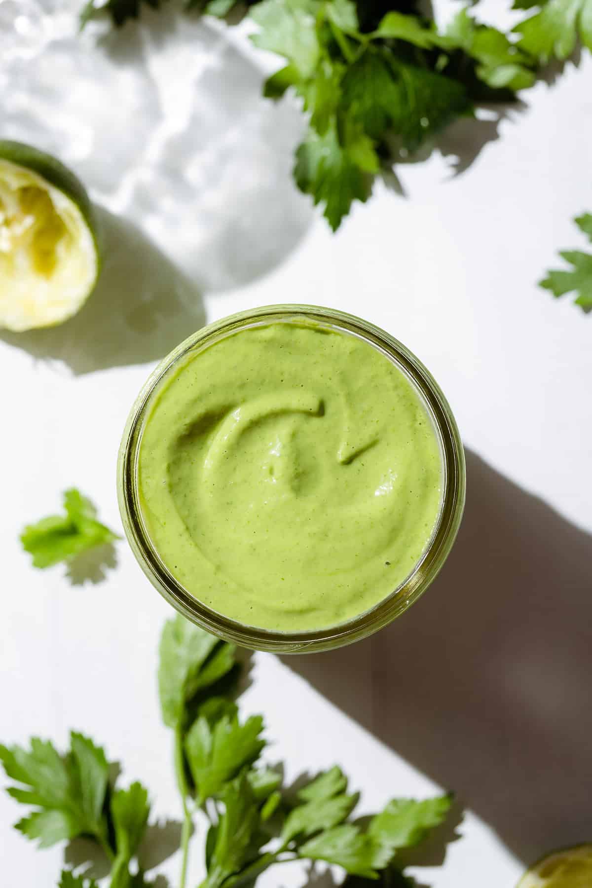 A mason jar with thick green dressing on a white background with fresh parsley around.