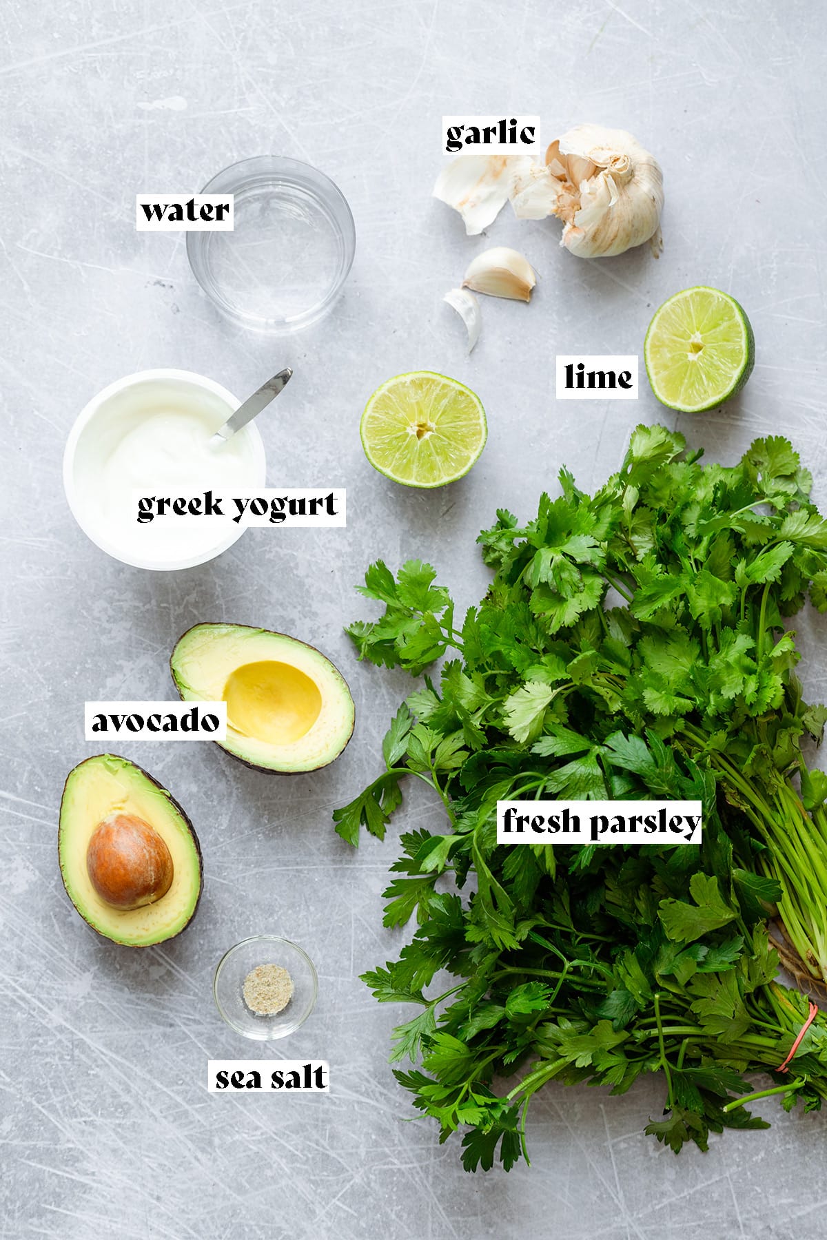 Parsley, avocado, lime, garlic, greek yogurt, and water laid out on a metal background.