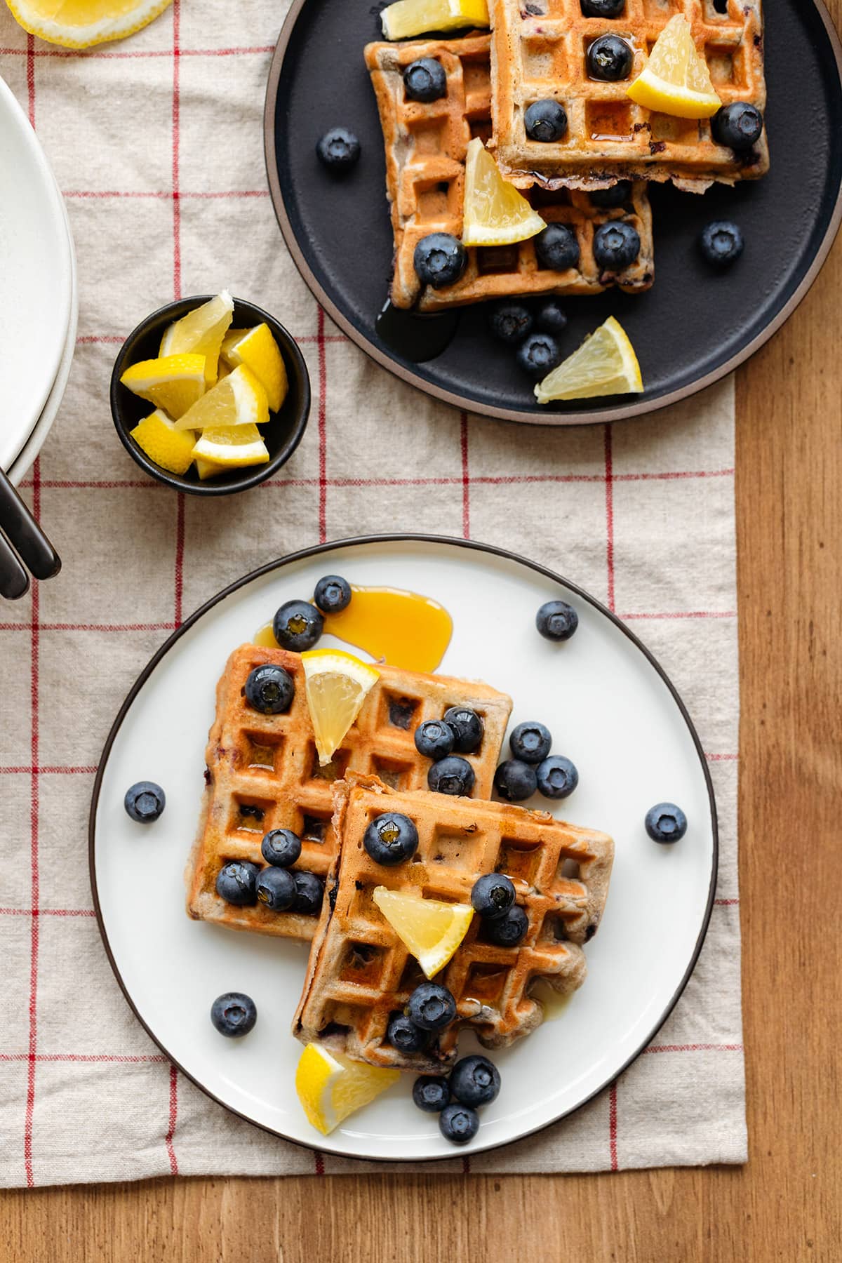 Waffles with blueberries and lemon slices on a white and black plates and a wooden table.