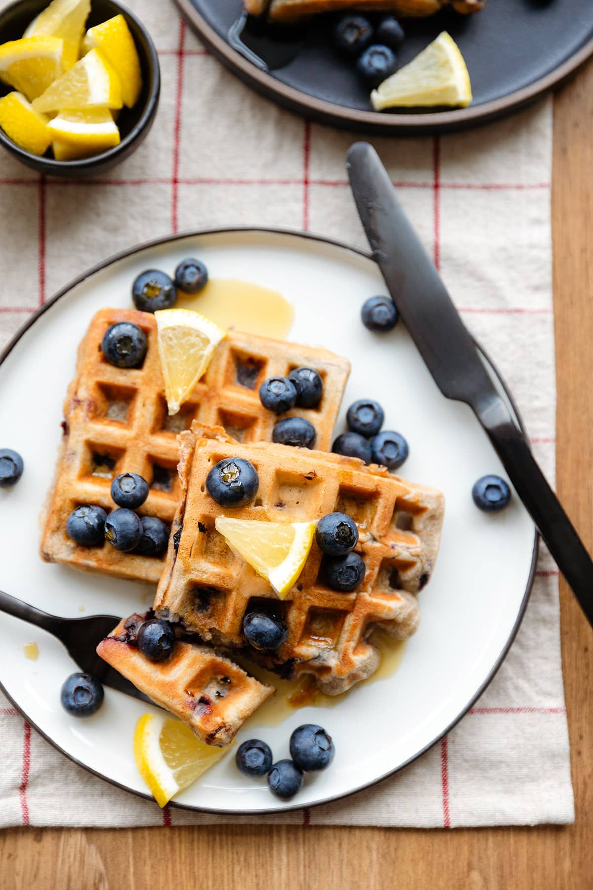 Waffles with blueberries and lemon slices on a white and black plates and a wooden table with fork and knife cutting in.