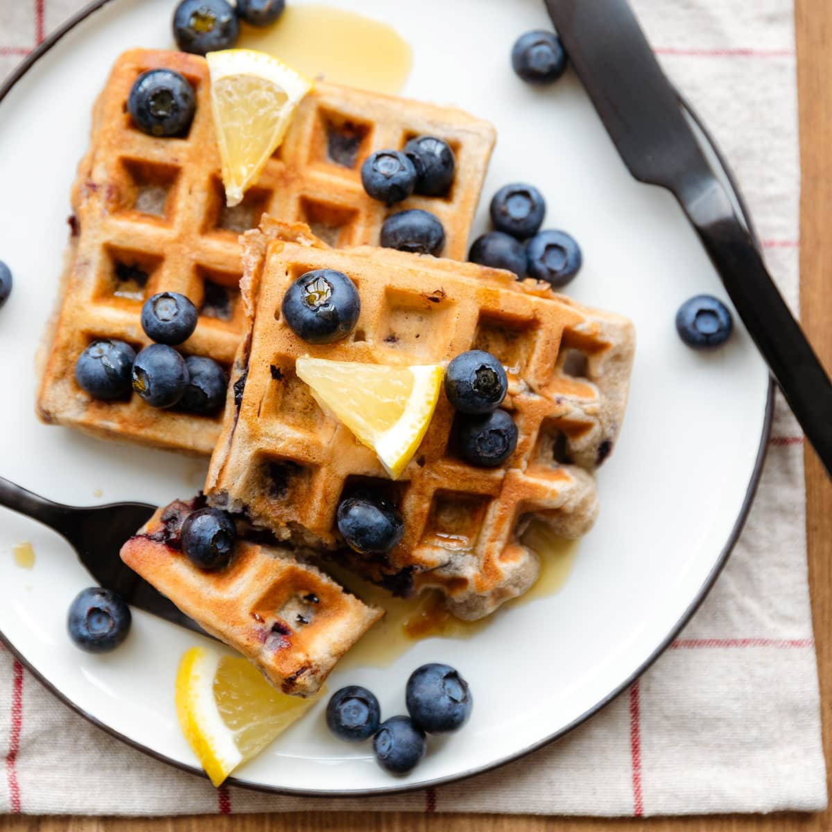 Waffles with blueberries and lemon slices on a white and black plates and a wooden table.