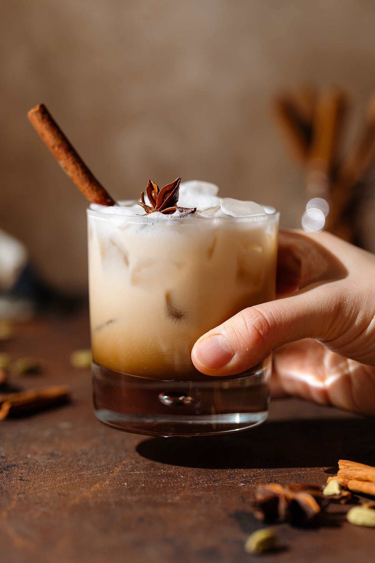A hand holding a White Russian cocktail in a double old fashioned glass garnished with a cinnamon stick and star anise on a wooden backdrop.