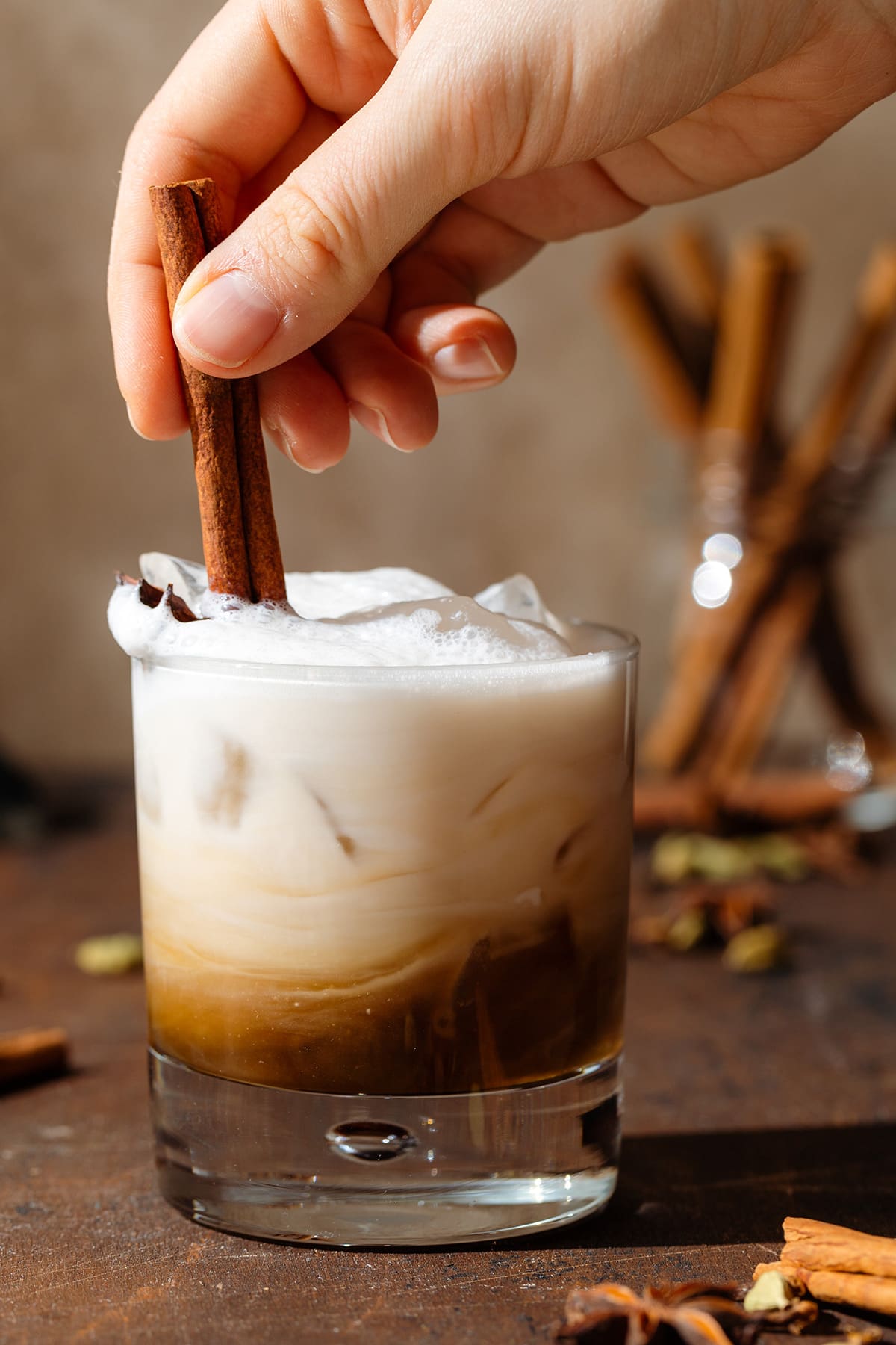 A hand mixing a white and brown-colored cocktail in a short glass together with a cinnamon stick.