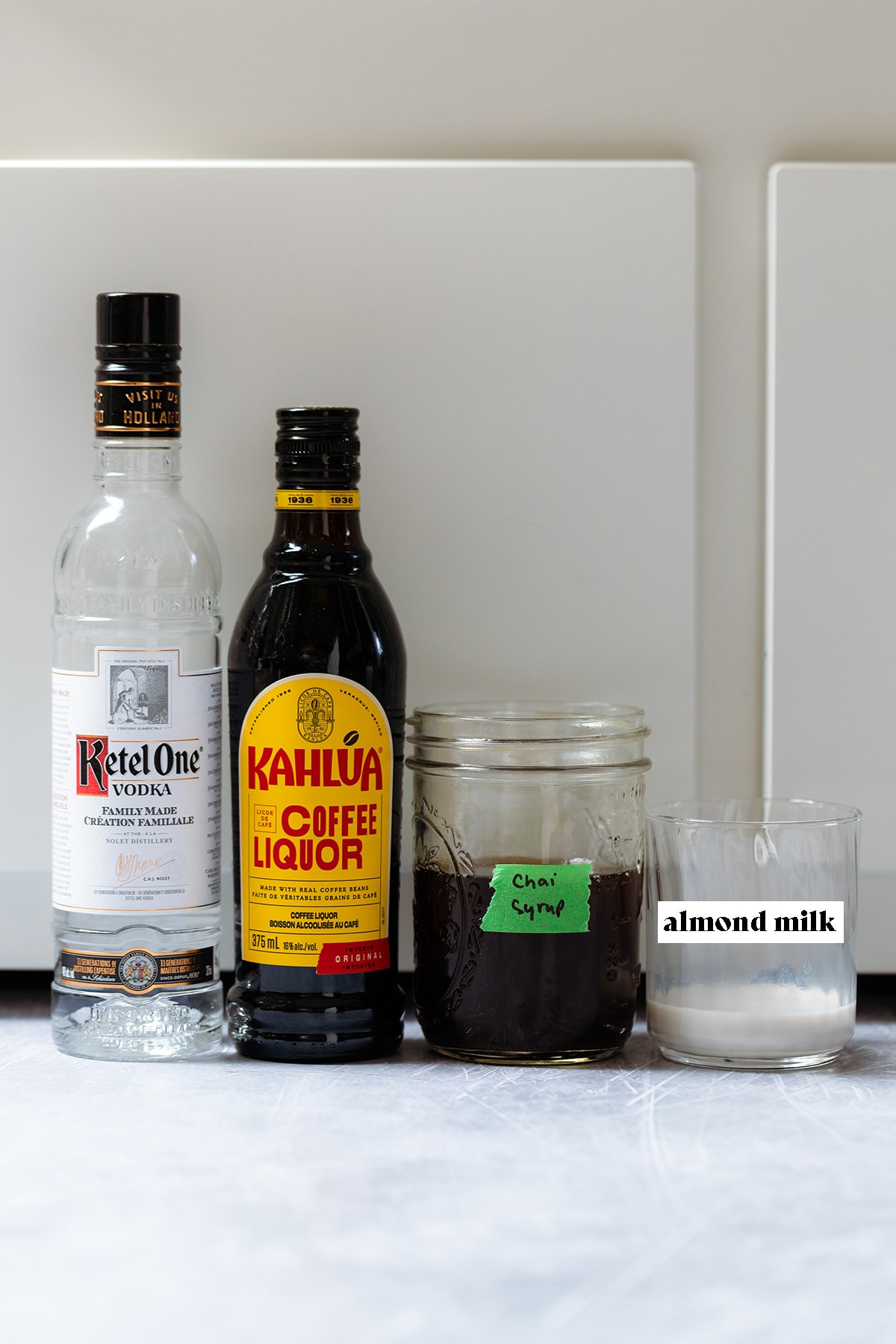 Vodka, Kahlua, chai syrup, and almond milk all on a white background.