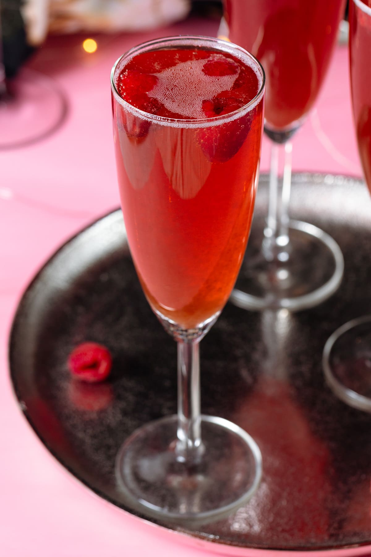 Pink Champagne cocktail with raspberries in flute glasses on a silver platter and pink and black background.