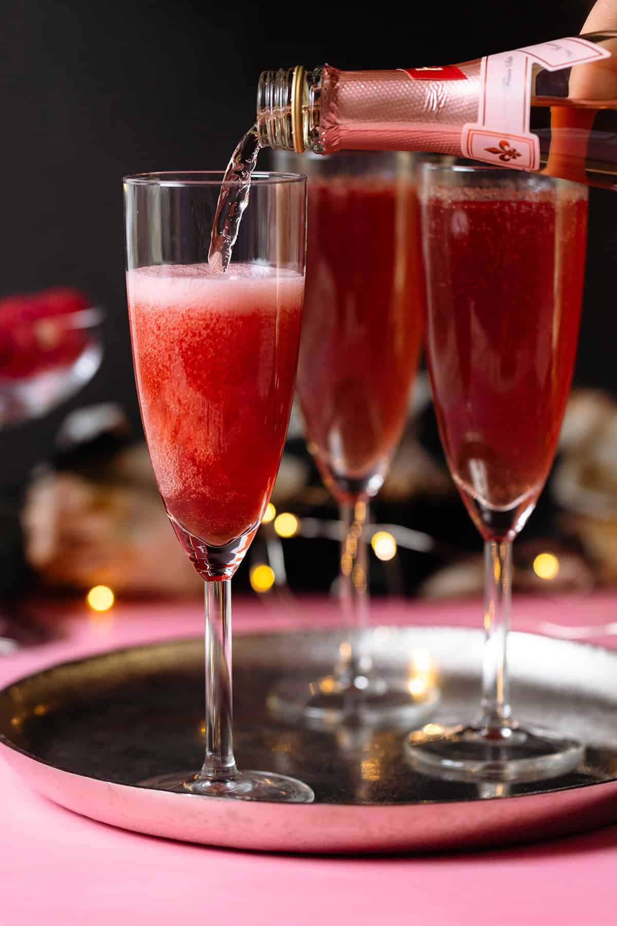 Pink Champagne cocktail being poured into flute glasses on a silver platter and pink and black background.