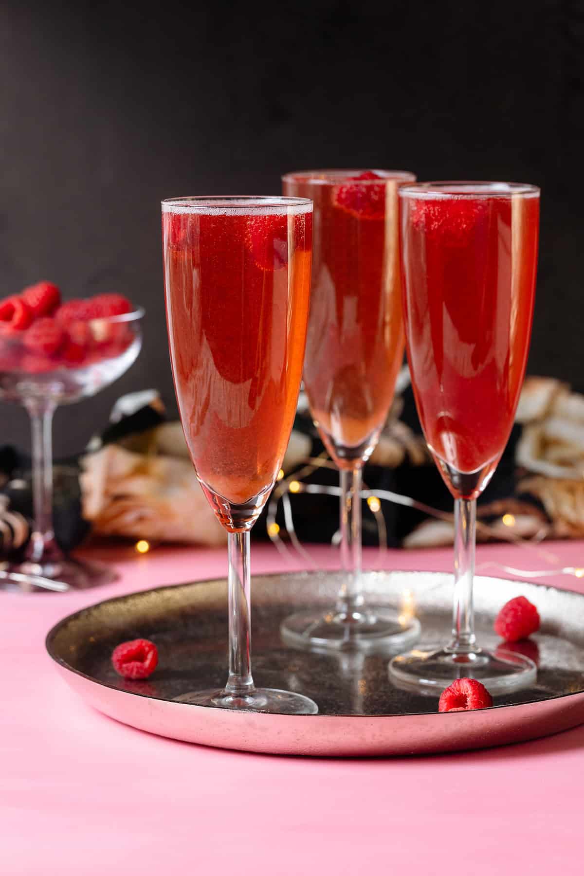 Pink Champagne cocktail with raspberries in flute glasses on a silver platter and pink and black background.