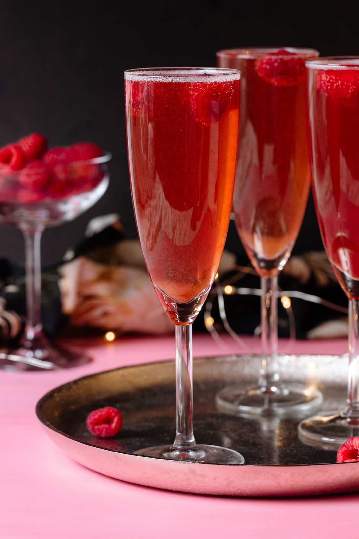 Pink Champagne cocktail with raspberries in flute glasses on a silver platter and pink and black background.